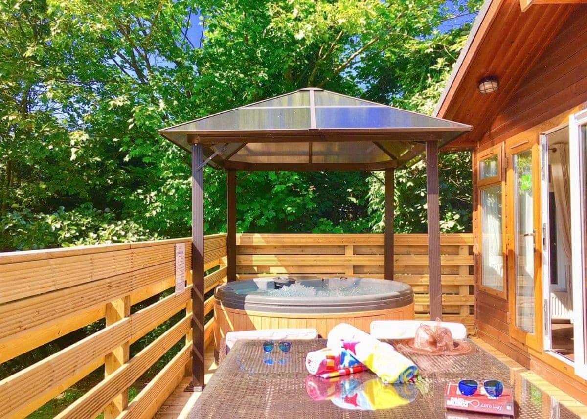A wooden lodge with an outdoor hot tub underneath a wooden gazebo. The lodge sits on a deck, with snacks and drinks set on a table, creating a perfect relaxation spot in a forest setting.