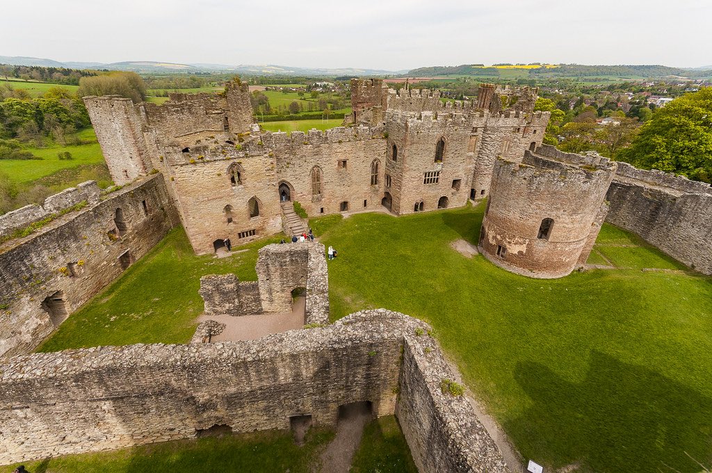 Aerial view of a large medieval castle ruins with stone walls, towers, and a grassy courtyard. The castle is surrounded by green countryside and rolling hills. In the distance, a small town and patchwork of fields can be seen. This appears to be Ludlow Castle in Shropshire, England, showcasing its expansive layout and picturesque setting.