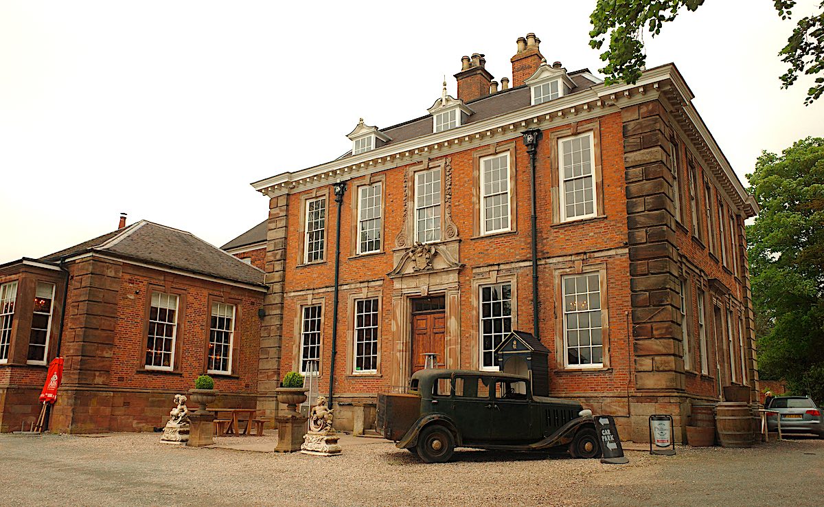 A large, historic manor-style building made of brick with tall windows and an ornate entrance. There is an old-fashioned car parked in front, adding to the historical atmosphere.