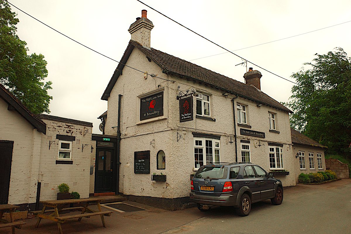 Another classic English pub with white walls and dark accents. The sign identifies it as "The Red Lion," and a small outdoor seating area is visible. A few parked cars are in front.
