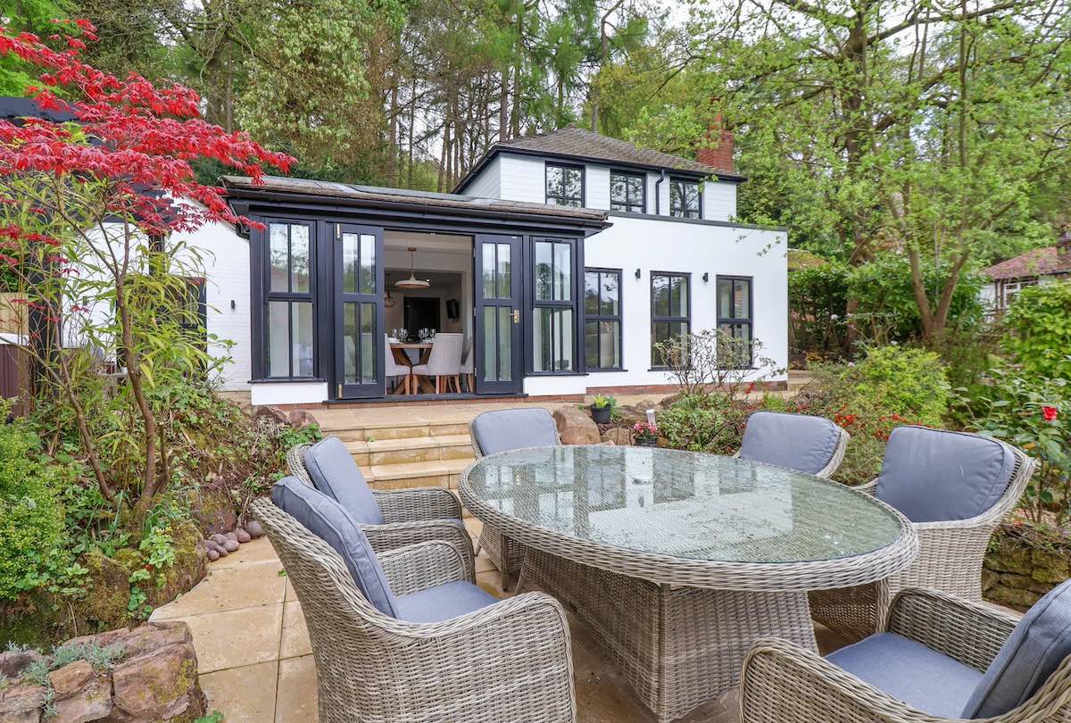 A stylish outdoor seating area featuring wicker chairs and a glass table set on a patio surrounded by lush plants and trees. The setting includes a modern black and white house with large windows in the background.