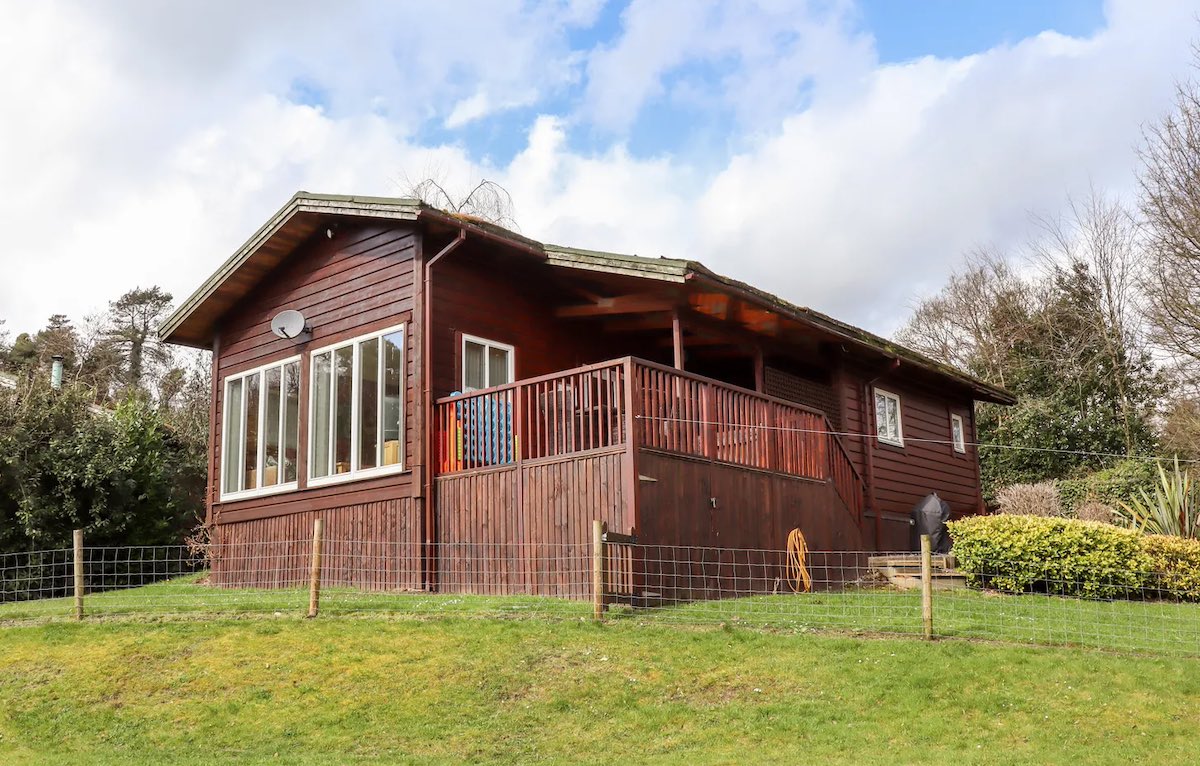 A cozy wooden cabin located in a peaceful, green forest setting. The cabin has a deck with outdoor seating and is enclosed by a simple wooden fence.