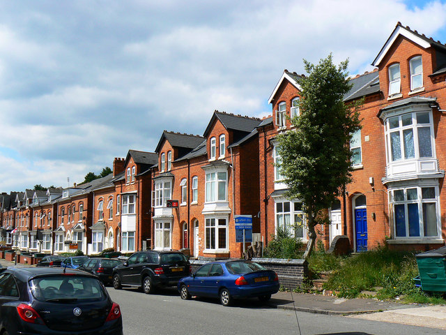 The image shows a row of tall red-brick terraced houses along a residential street. The homes feature bay windows, arched doorways, and decorative white trim. A mix of cars is parked along the curb, and one of the houses has a "For Sale" sign in front. A small tree grows near the sidewalk, and some greenery is visible in front of a few homes. The sky is partly cloudy, and the overall scene conveys a typical urban neighborhood with Victorian-style architecture.
