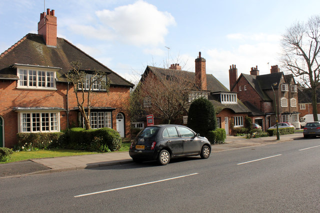 The image shows a row of detached brick houses along a suburban street. The homes have sloped, dark-tiled roofs, prominent chimneys, and well-maintained front gardens with shrubs and small trees. A black car is parked on the roadside, and there is a "For Sale" sign visible in front of one house. The sky is partly cloudy, and the trees in the background are bare, suggesting early spring or late winter. The scene feels peaceful and residential, with a quiet neighborhood atmosphere.