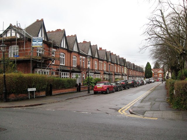 A residential street lined with red brick terraced houses. The houses have a distinct architectural style with gabled roofs and bay windows. There's scaffolding on one of the houses, indicating renovation work. Cars are parked along the street, and bare trees can be seen on the right side. The street appears to be in a typical British neighborhood.