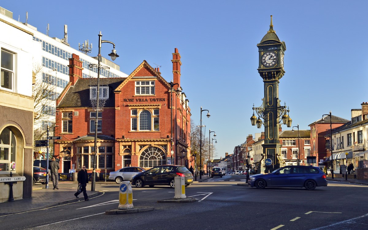 A street scene in a town center, dominated by a large ornate clock tower in the foreground. The clock tower is dark green and features multiple clock faces and decorative elements. To the left is a red brick building with a sign reading "Rose Villa Tavern". The street is busy with cars and pedestrians, and other shops and buildings line the road, giving a sense of a bustling small town or city center.