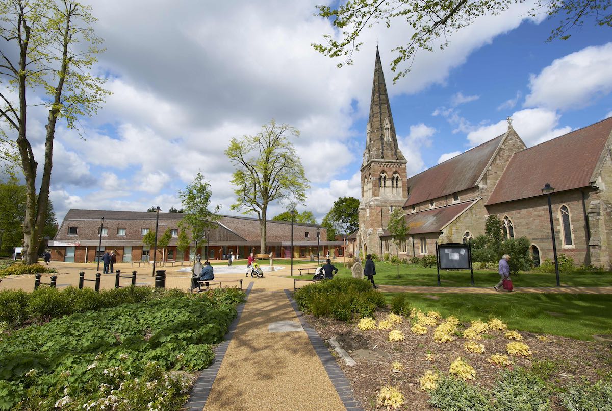 A picturesque town square with a large stone church as the focal point. The church features a tall, ornate spire. In the foreground, there's a well-maintained public space with benches, landscaped gardens, and paved walkways. Modern buildings blend with the historic church, creating a harmonious town center. People can be seen walking and relaxing in the area on a partly cloudy day.