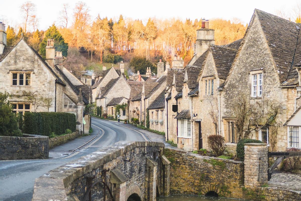A winding road through a Cotswold village with stone cottages and a small bridge over a stream.
