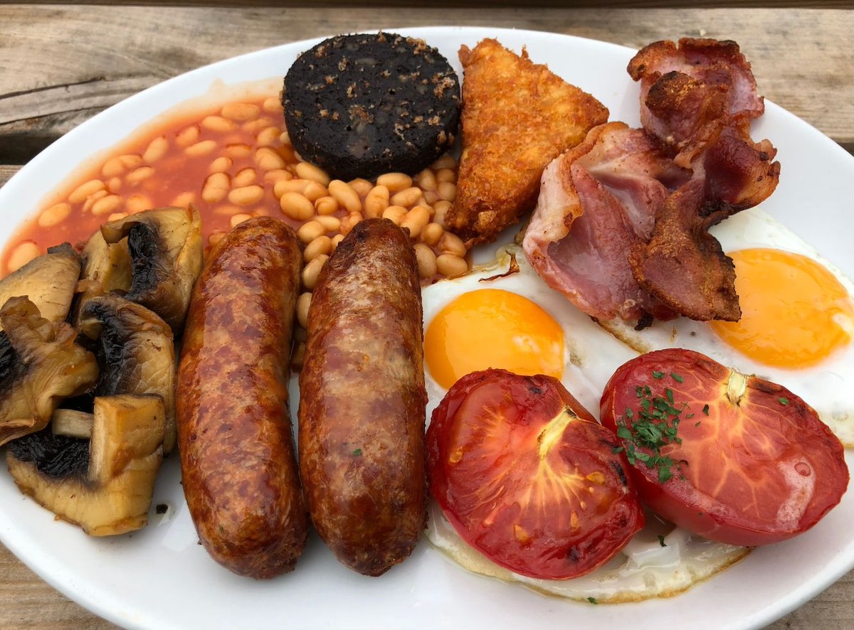 A full English breakfast on a white plate, including fried eggs, sausages, bacon, grilled tomatoes, mushrooms, baked beans, hash browns, and what appears to be black pudding.