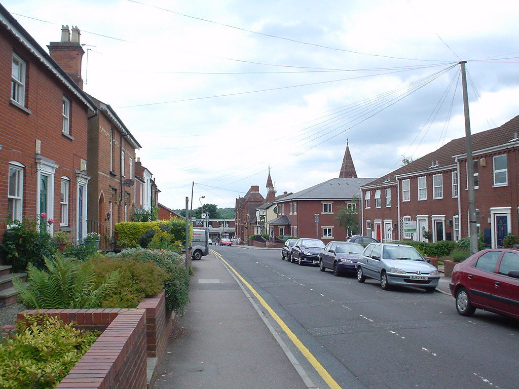 A street view of a typical English town. The road is lined with brick houses on both sides, some with small front gardens. Parked cars are visible along the street. In the distance, church spires can be seen rising above the rooftops. Overhead power lines crisscross the sky. The scene depicts a mix of residential and commercial buildings, with double yellow lines on the road indicating parking restrictions.