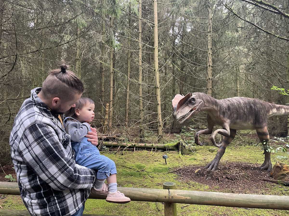 A father holding his young child in a forested area, both looking towards a nearby dinosaur model of a Velociraptor. The dinosaur is posed as if in motion, creating a sense of interaction and curiosity.