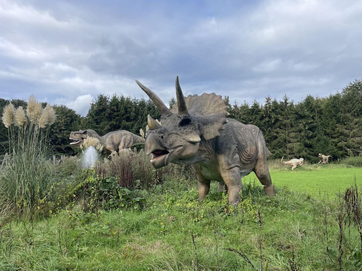 Life-sized model of a Triceratops dinosaur in a grassy, natural setting, with trees and a cloudy sky in the background. Other dinosaur models are partially visible, adding depth to the scene.