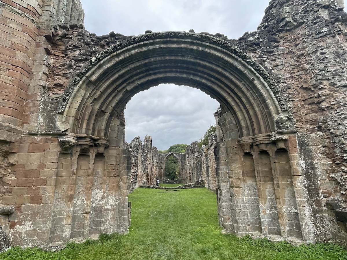 Stone archway from the ruins of an ancient abbey, showcasing intricate stonework against a backdrop of grass and scattered ruins beneath a cloudy sky.