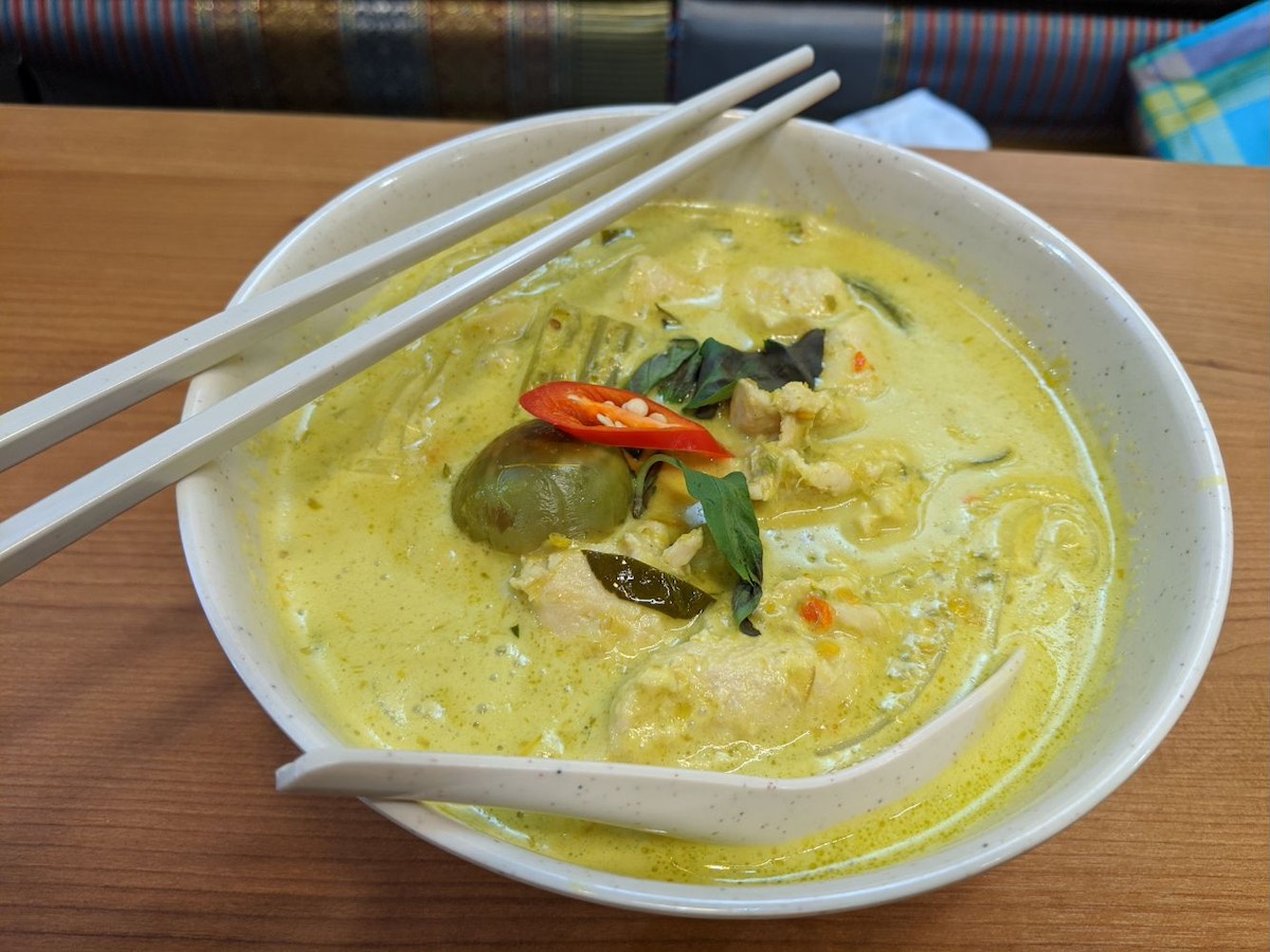 A bowl of yellow curry soup with visible pieces of meat and vegetables, garnished with a red chili pepper and herbs. Chopsticks rest on the side of the bowl.