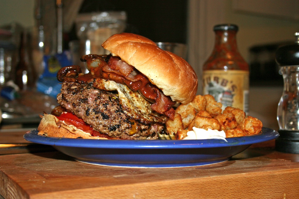 A hearty homemade burger with bacon, a beef patty with grill marks, and other toppings on a golden bun. It's served on a blue plate with what look like seasoned potato puffs or tater tots. A bottle of sauce and salt shaker are visible in the background on what appears to be a wooden kitchen counter.