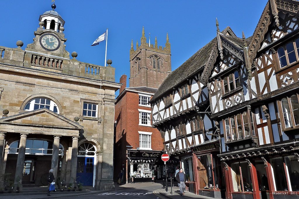 A historic town center featuring a stone building with a clock tower on the left, and traditional half-timbered Tudor-style buildings on the right. A church spire is visible in the background, and the scene shows a clear blue sky.
