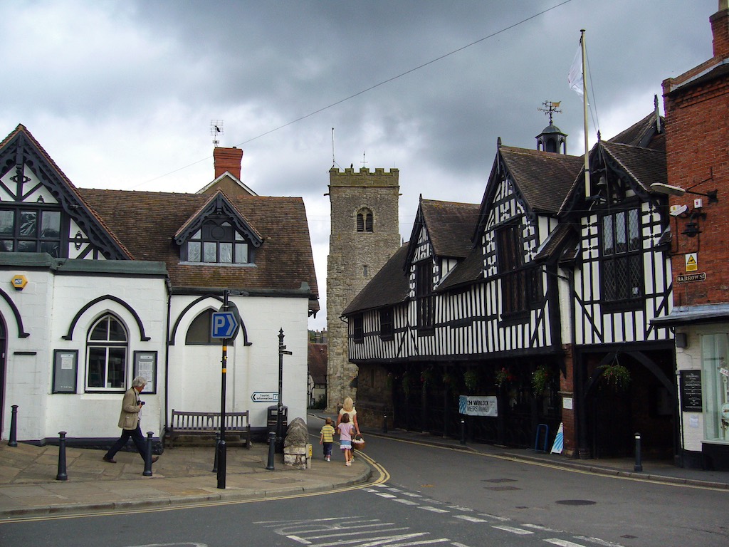 A picturesque street scene in a small town, featuring Tudor-style half-timbered buildings with black and white facades. A stone church tower is visible in the background, and a few people walk along the street.