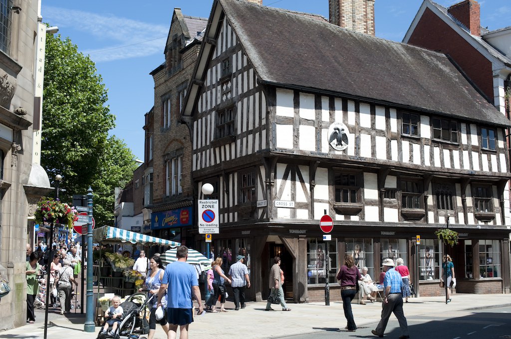 A busy street in a historic town center with a prominent half-timbered Tudor-style building on the right. People are walking on the sidewalk, including someone pushing a baby stroller. Street signs and shops are visible, with trees and buildings in the background.
