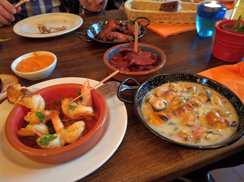 A spread of Spanish tapas or appetizers on a wooden table. The dishes include garlic shrimp in a red ceramic bowl, a creamy seafood dish that appears to be a shellfish stew in a small black pan, what looks like cured meat in a small bowl, and various other small plates and condiments. A blue candle holder and bread basket are visible in the background.