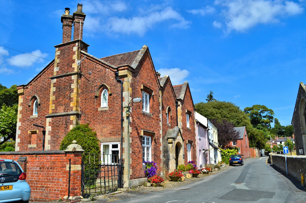 A quaint English street with red brick Victorian-style houses. Colorful flower planters line the road, and a blue car is parked near a white-painted gate. The scene is bathed in bright blue sky and sunshine.