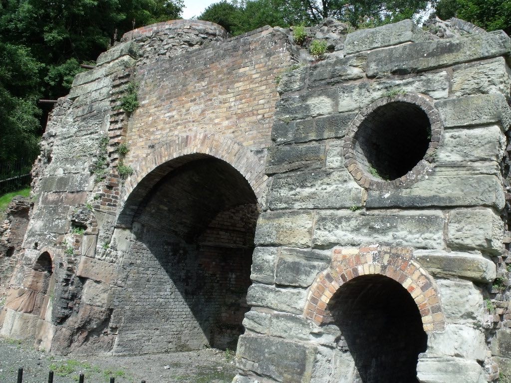 Ancient stone structure with multiple arched openings of different sizes. The ruins appear to be part of an old industrial building, possibly a kiln or furnace, with a large central arch and smaller circular openings. The structure is made of weathered stone and brick masonry with some vegetation growing on it. Trees are visible in the background.