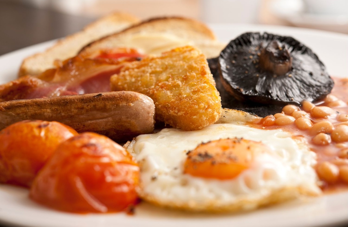 Close-up of a traditional full English breakfast on a white plate featuring fried eggs, bacon, sausage, hash brown, grilled tomatoes, beans, mushroom, and toast. The dish shows perfectly cooked components with vibrant colours and textures.