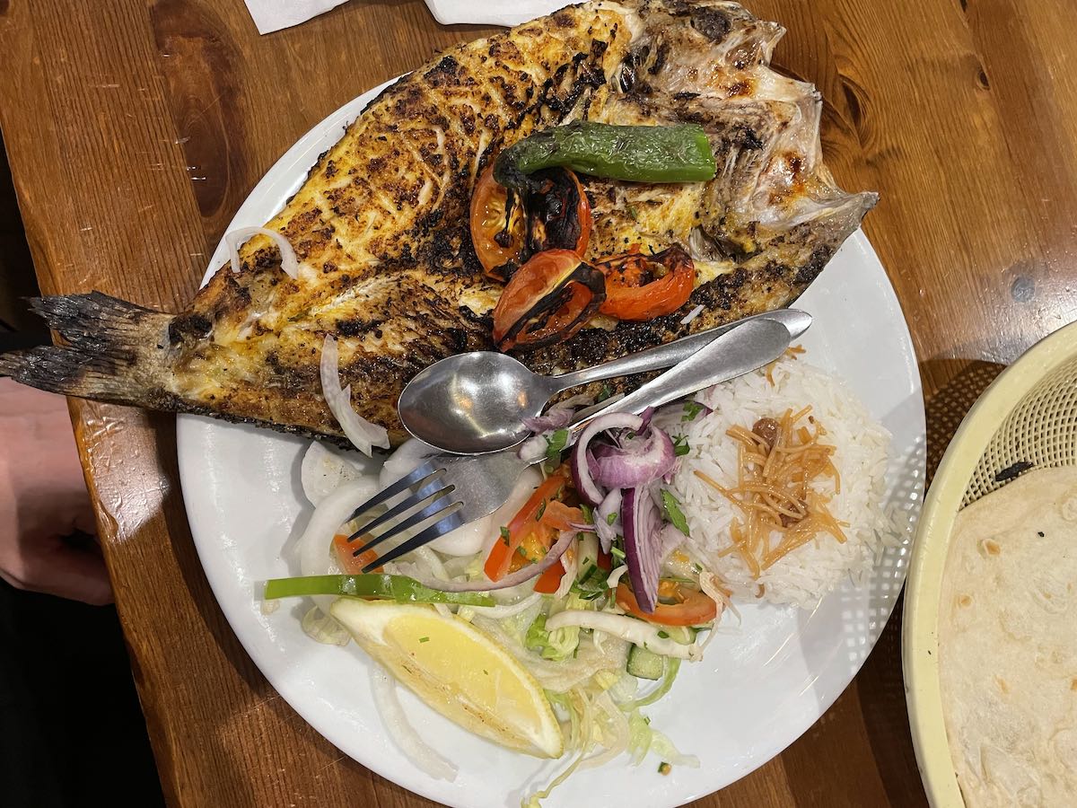 A grilled whole fish served on a white plate with roasted bell peppers and tomatoes on top. The plate also includes white rice with vermicelli, a fresh vegetable salad, and a lemon wedge. Cutlery is visible on the plate, and the dish is presented on a wooden table.