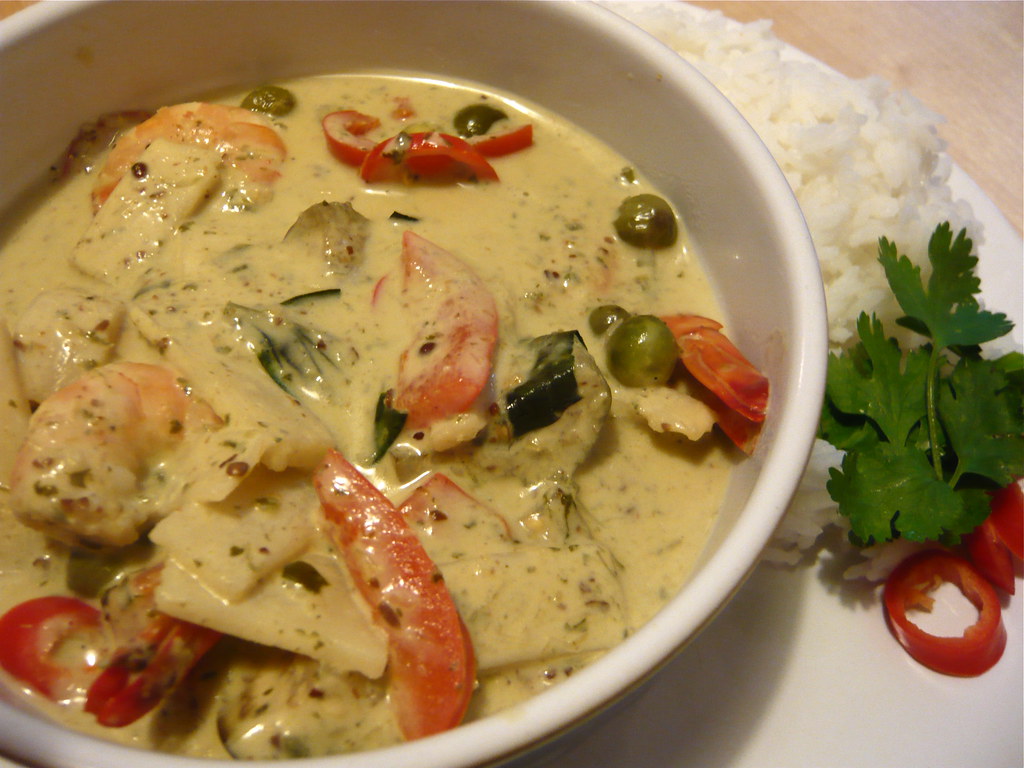 A creamy Thai green curry served in a white bowl, featuring chunks of chicken, sliced red bell peppers, green peas, and what appears to be zucchini in a pale yellow-green coconut sauce. The curry is accompanied by a side of white rice, garnished with fresh cilantro leaves and thin slices of red chili pepper for color and heat.