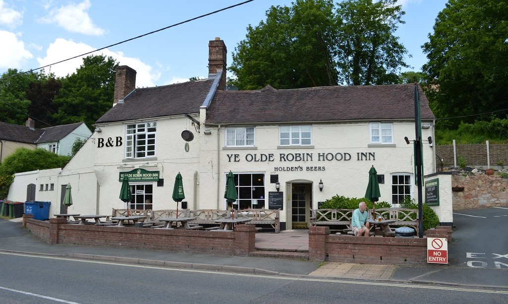 "Ye Olde Robin Hood Inn," a white-painted traditional pub with "B&B" signage and "Holden's Beers" on display. The building has an outdoor seating area with wooden benches and green umbrellas, brick low walls around the patio, and is situated along a road with trees in the background.