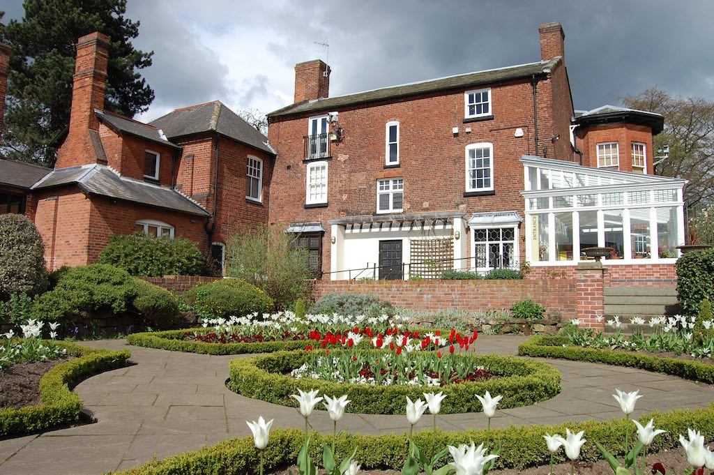 A large red brick manor house with multiple chimneys and a conservatory extension on the right side. The house has white-framed windows and multiple wings. In the foreground is a formal garden with neatly trimmed box hedges arranged in geometric patterns, stone pathways, and flowering tulips in red and white. The garden is well-maintained with various ornamental plantings against a backdrop of dark cloudy skies.