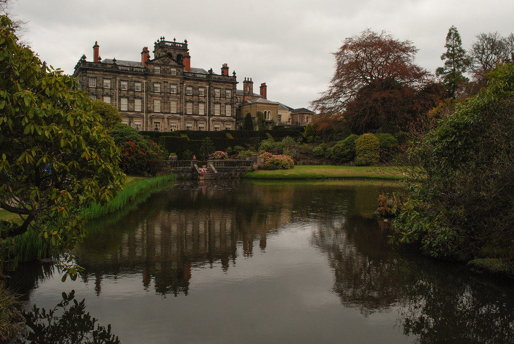 A grand Victorian mansion with ornate architecture reflected in a calm pond surrounded by lush greenery, with various bushes and trees adding a sense of tranquility to the landscape. The sky is overcast, adding to the garden’s serene atmosphere.