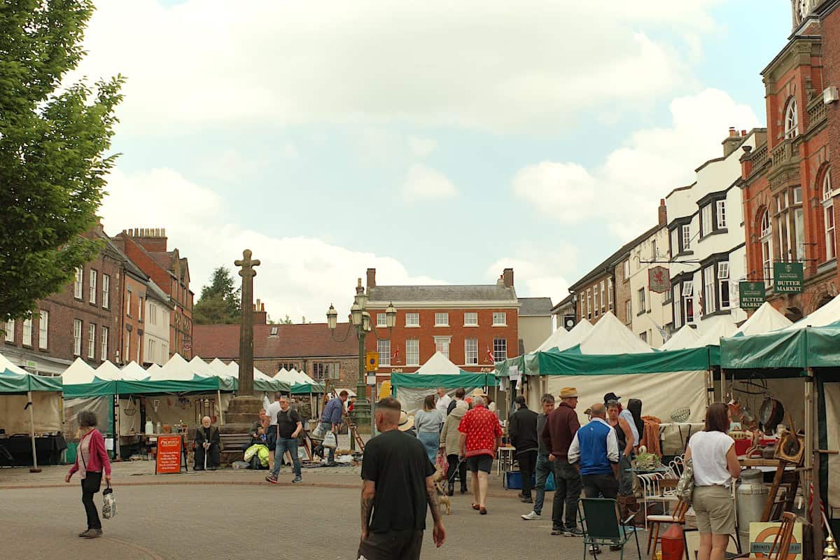 a large market square in the centre of an old town, with white and green marquee tents lined around the perimeter selling goods, while lots of people walk around and browse the markets. In the background are some old edwardian houses and in the centre is an old memorial statue.