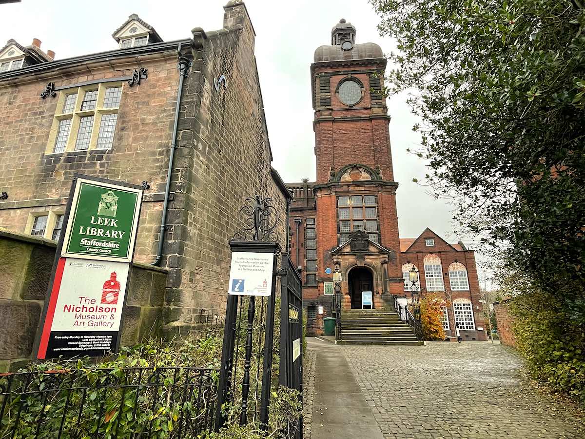 Historic Leek Library building and Nicholson Museum & Art Gallery in Staffordshire. The building features a prominent red brick clock tower with stone trim, stone steps leading to the entrance, and signs indicating both the library and museum. The building combines Victorian Gothic architecture with local sandstone and brick construction. A cobbled path leads up to the entrance, with ornate iron gates and landscaping in the foreground.