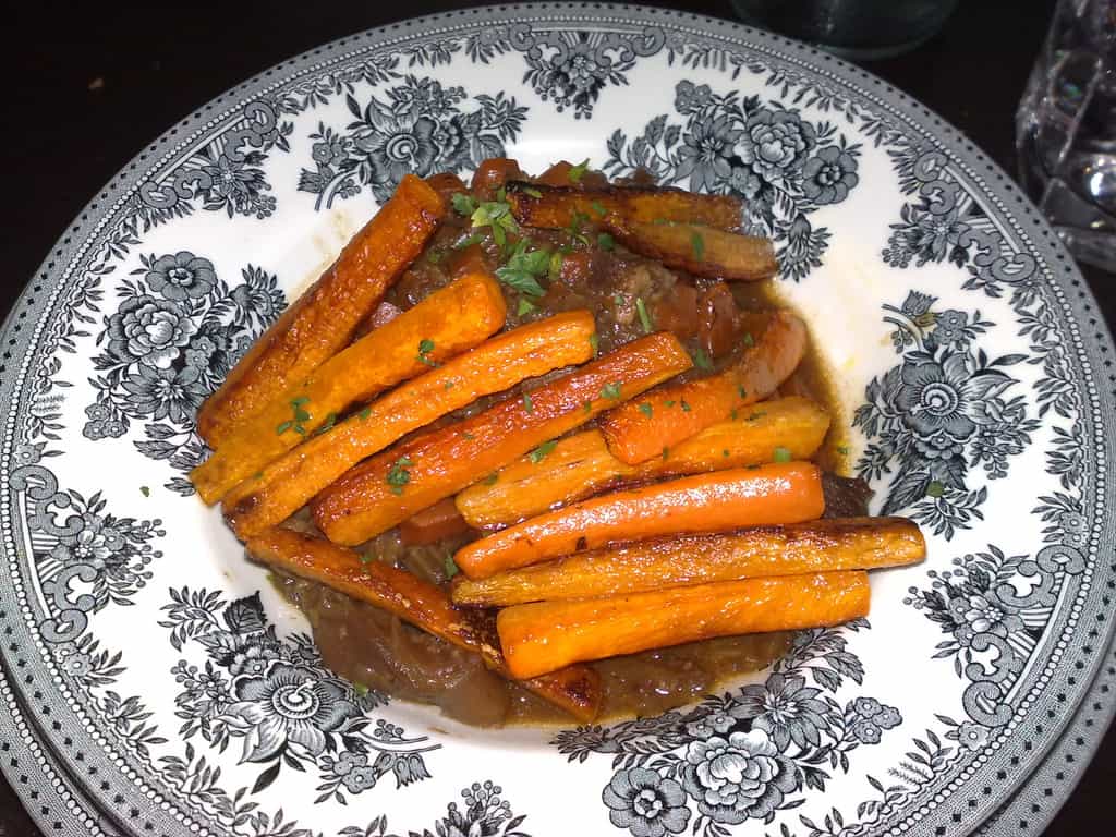 Glazed carrots arranged on an ornate white plate with black floral transferware pattern. The carrots appear honey-glazed and garnished with fresh herbs, presented in an elegant dining setting.