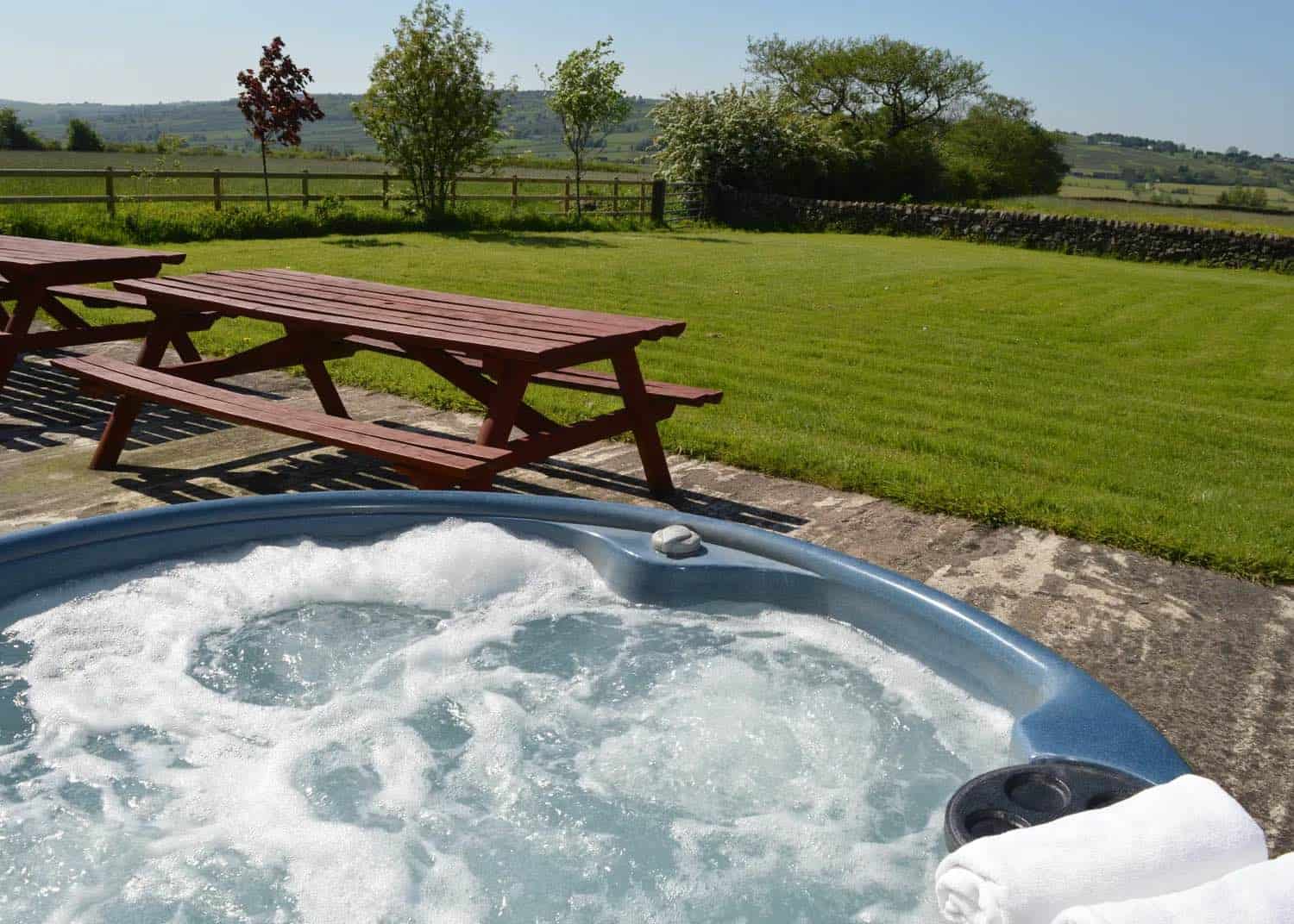 Active hot tub with bubbling water and foam, set on a stone patio beside wooden picnic benches, overlooking expansive green fields and countryside with scattered trees and hedgerows.