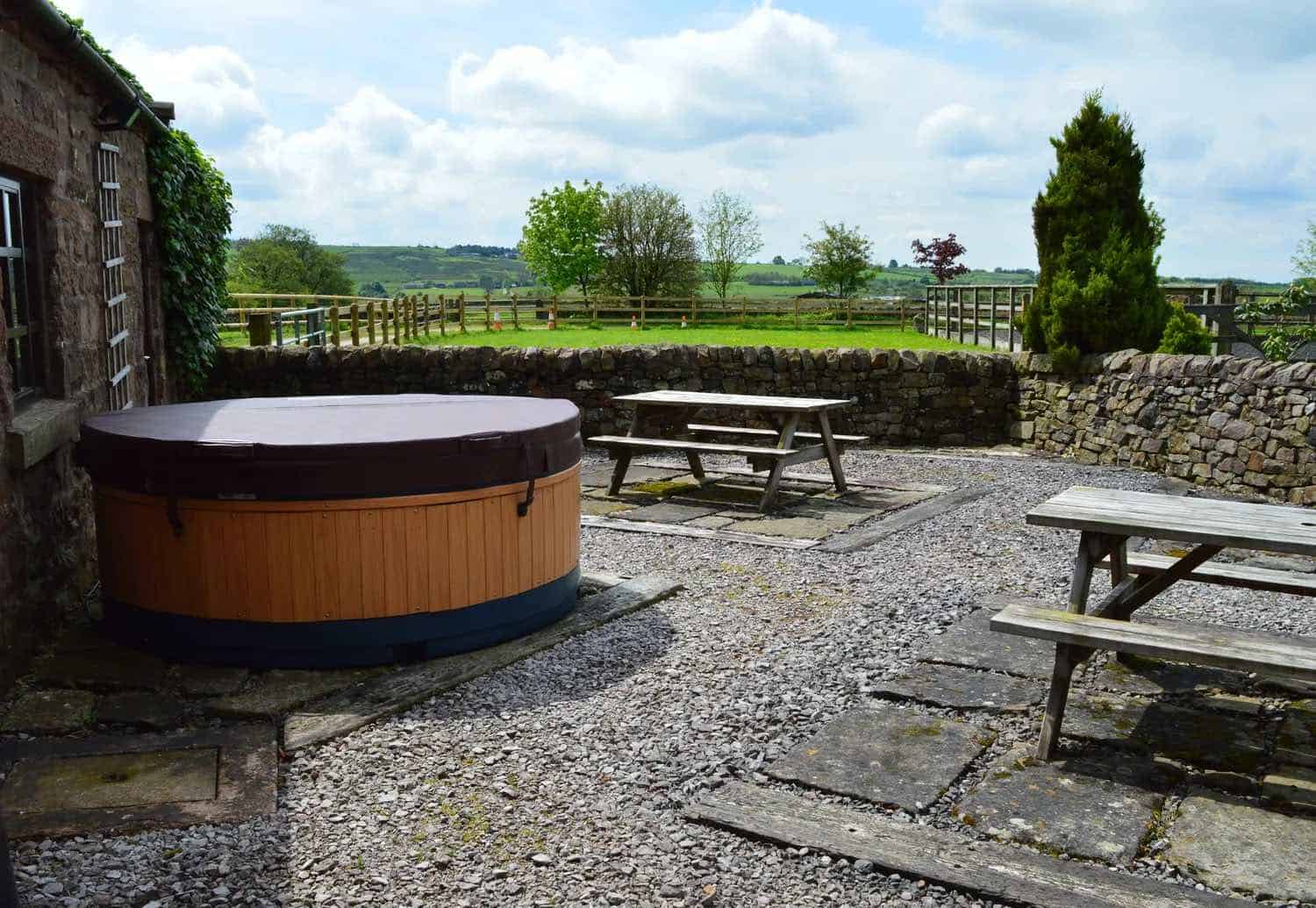 Circular wooden hot tub with dark cover positioned on a gravel patio area, surrounded by weathered wooden picnic tables and benches, with traditional dry stone walls and rural English countryside views in the background.