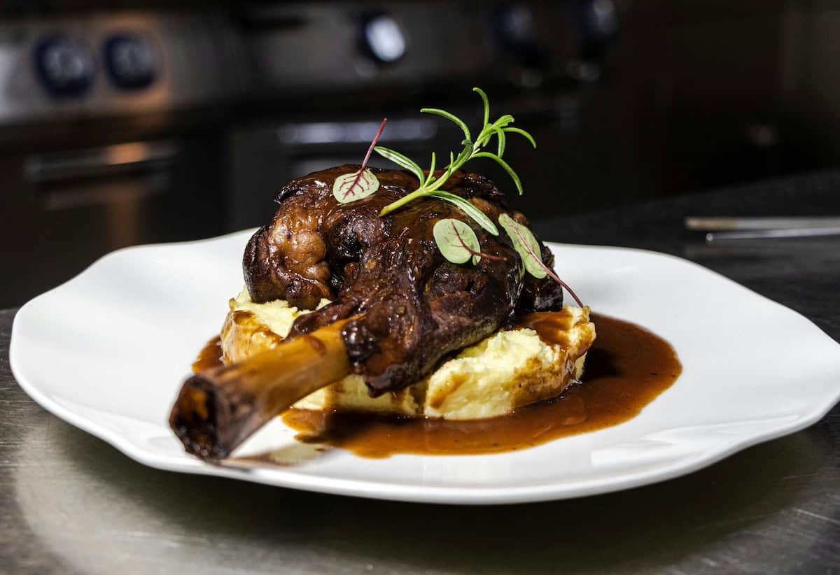 A gourmet lamb dish featuring a rack of lamb with rich brown gravy, served on a bed of golden Yorkshire pudding. The plate is garnished with fresh rosemary sprigs and radish slices, photographed in a professional kitchen setting.