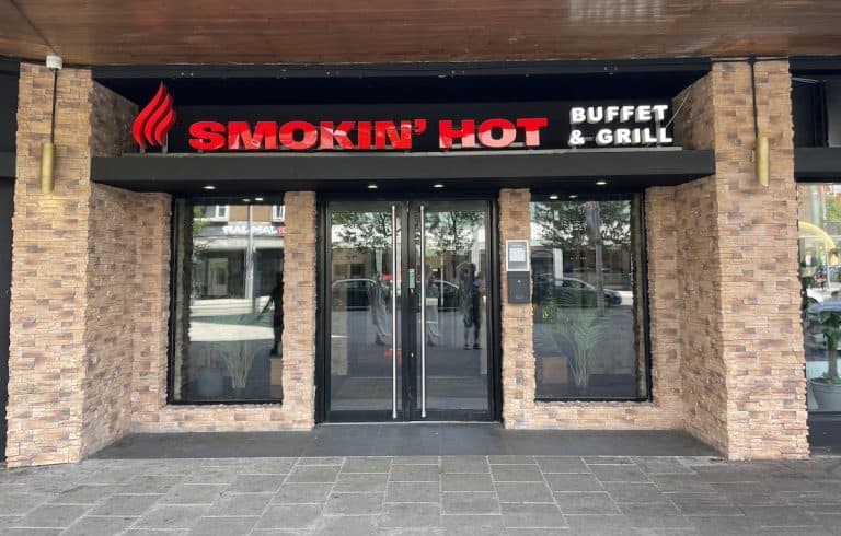 The exterior storefront of "Smokin' Hot Buffet & Grill" restaurant, featuring a brick facade with large glass windows and doors, and red signage with a flame logo.