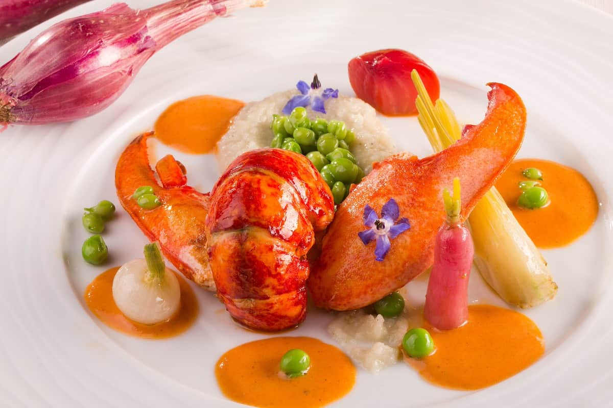 An artistically plated lobster dish with vibrant orange sauce dots around the plate. The lobster is garnished with fresh peas, colorful vegetables, edible flowers, and decorative red onion brushstrokes, creating an elegant fine dining presentation.
