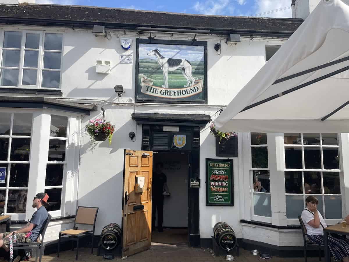 The Greyhound Inn pub, a white-painted traditional English pub with black trim and a large painted sign featuring a greyhound dog. The building has hanging flower baskets, wooden beer barrels outside, and outdoor seating. A 'Multi-Award Winning Venue' sign is visible by the entrance.