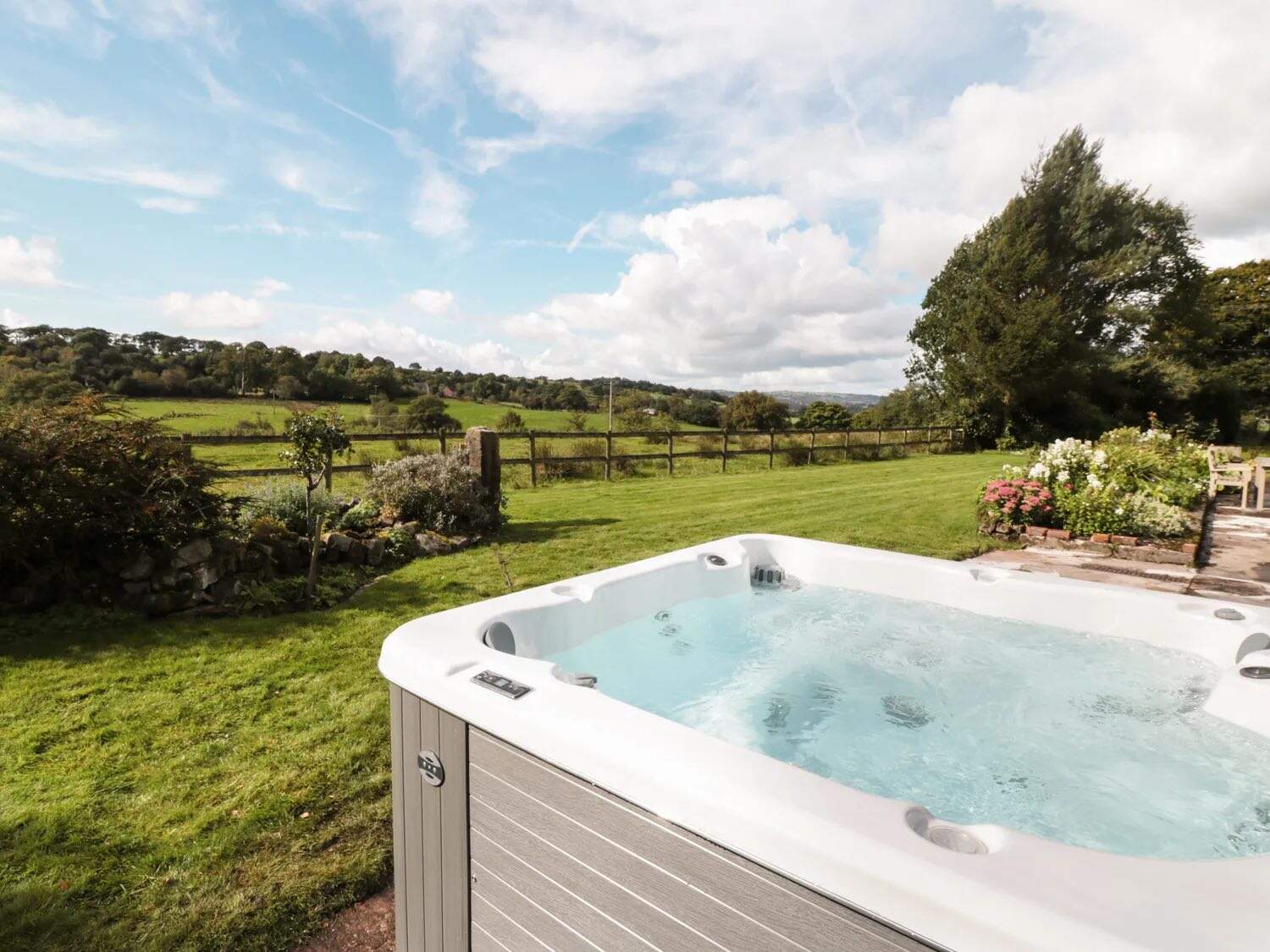 Large white hot tub positioned on manicured lawn with panoramic views across green fields and countryside, featuring built-in seating and modern spa equipment, with flowering garden borders nearby.