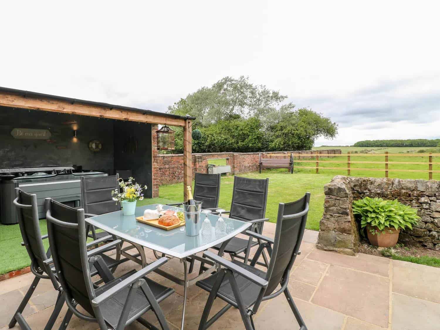 Modern outdoor dining area with glass-top table and six charcoal gray chairs on a stone patio, featuring an outdoor kitchen with stainless steel appliances under a wooden pergola, overlooking green countryside with rolling hills and traditional stone walls.