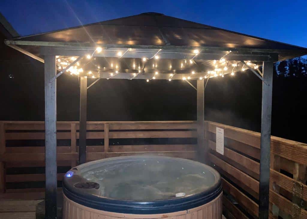 Evening view of circular hot tub under wooden gazebo structure illuminated by warm string lights against a deep blue twilight sky, creating an intimate outdoor relaxation space.