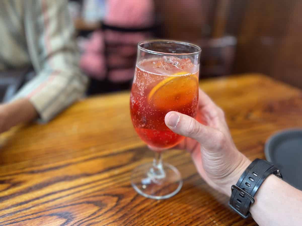 A hand wearing a black fitness watch holds a wine glass containing a reddish-orange cocktail garnished with an orange slice, set against the warm wooden interior of a pub or restaurant.