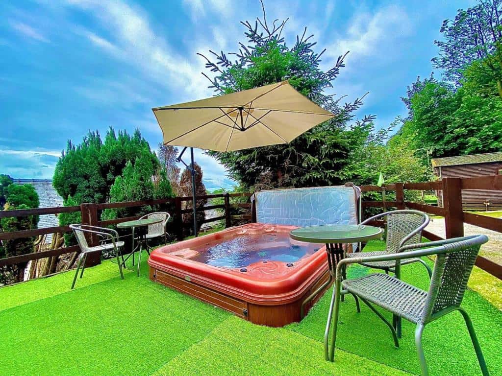 Square red hot tub on artificial grass decking, surrounded by wicker outdoor furniture and shaded by a large cream parasol, with rural countryside and residential buildings visible beyond wooden fencing.