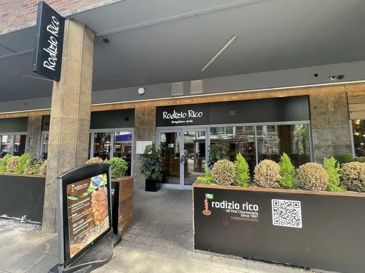 Rodizio Rico Brazilian Grill restaurant entrance in a modern shopping center or mall. The restaurant features dark signage, glass doors, and decorative planters with greenery. A promotional sign advertising churrascaria is visible outside the entrance.