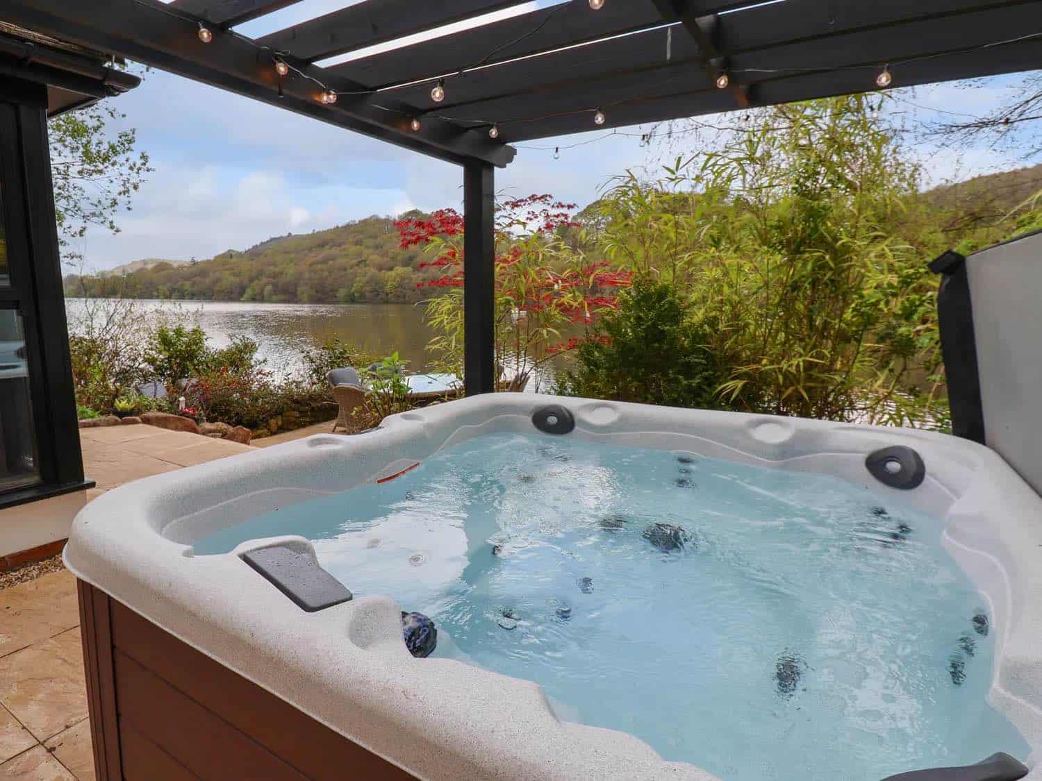 Lakeside hot tub positioned under a modern black pergola with string lights, featuring a scenic view across a lake toward wooded hills, with landscaped gardens and outdoor seating visible on the surrounding patio.