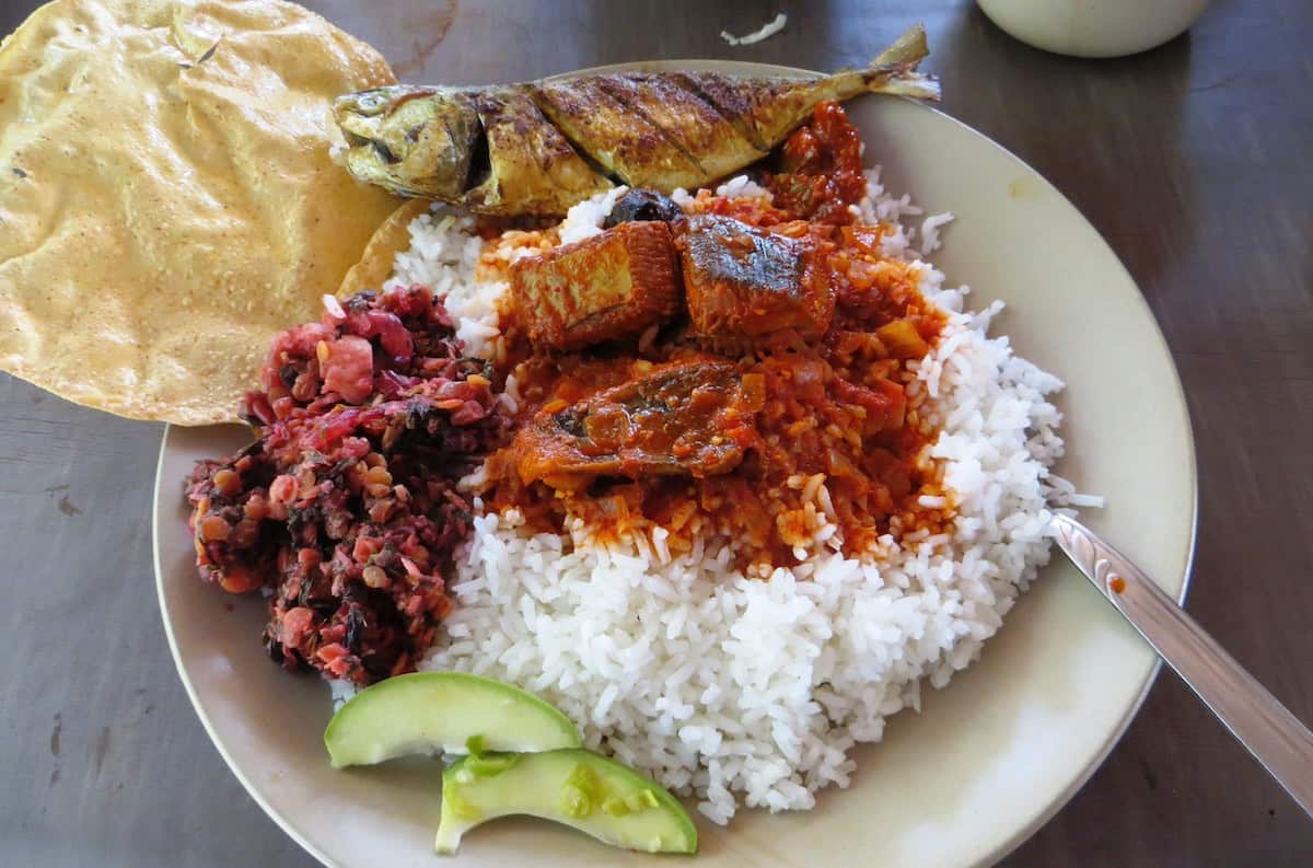 A traditional Sri Lankan rice and curry plate featuring white rice with various curries including what appears to be fish curry and a dark vegetable curry. The meal is accompanied by papadum, lime wedges, and served on a simple white plate.