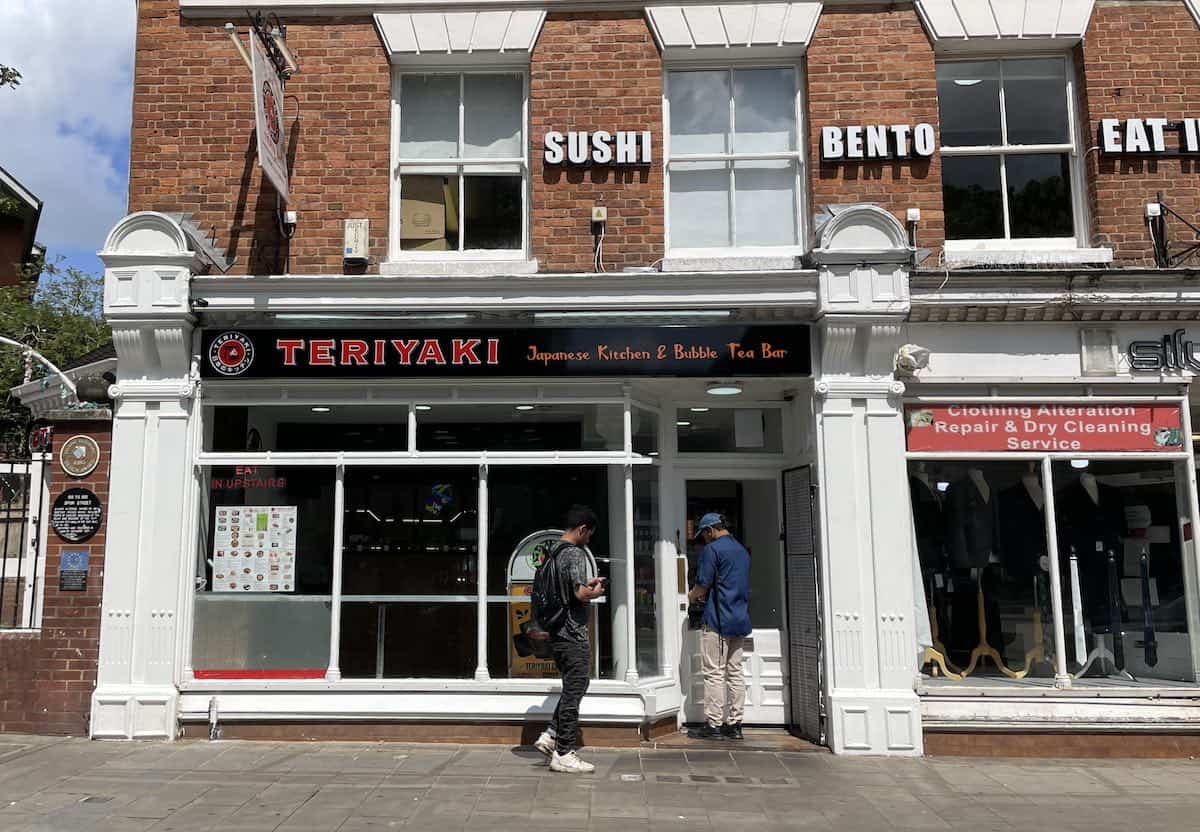 Teriyaki Japanese Kitchen & Bubble Tea Bar, a modern restaurant with red signage on a brick building. The storefront has large windows and white trim. Signs for 'SUSHI', 'BENTO', and 'EAT' are visible in the upper windows. People can be seen walking past on the street.