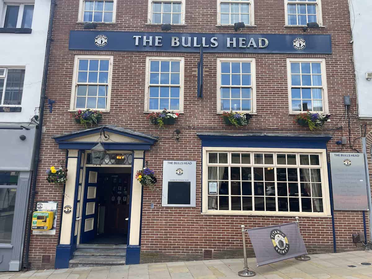 The Bulls Head, a traditional English pub housed in a red brick Georgian-style building with blue trim and signage. The pub features sash windows with decorative flower boxes, a classical entrance with triangular pediment, and large ground-floor windows. A pub sign and promotional board are visible on the street level.
